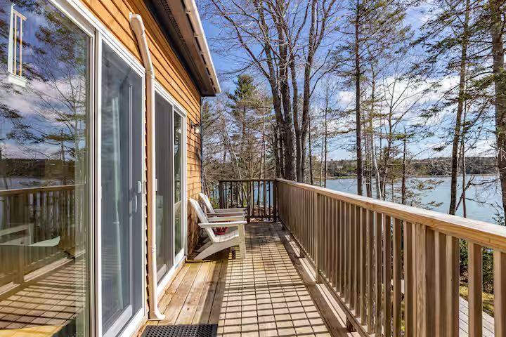 Wooden deck with railing beside a lakeside cabin, with sliding glass doors and chairs in the sunlight