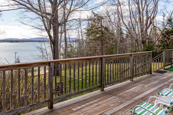 Wooden deck overlooking a lake, with railing, bare trees, and two striped lounge chairs