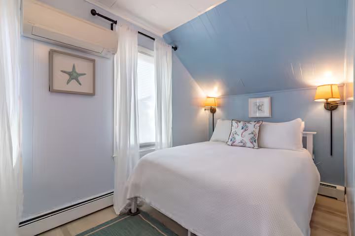 Bright coastal bedroom with a white bed, blue sloped ceiling, and wall sconces