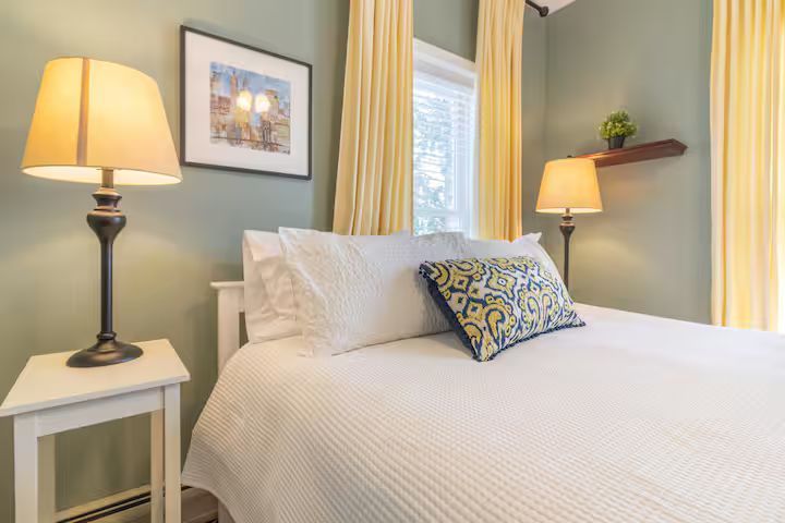 Bright bedroom with a white bed, bedside lamps, soft green walls, and yellow curtains by a window