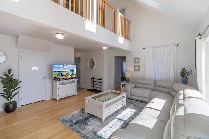 Bright living room with gray sofas, wood floors, TV console, coffee table, and loft railing above