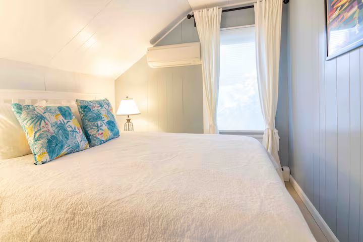 Bright bedroom with a white bed, blue patterned pillows, and a window with curtains and a lamp