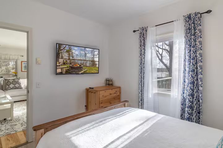 Bright bedroom with white walls, bed, wooden dresser, wall TV, and patterned curtains by the window