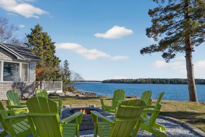 Green Adirondack chairs overlook a blue lake beside a house on a sunny day
