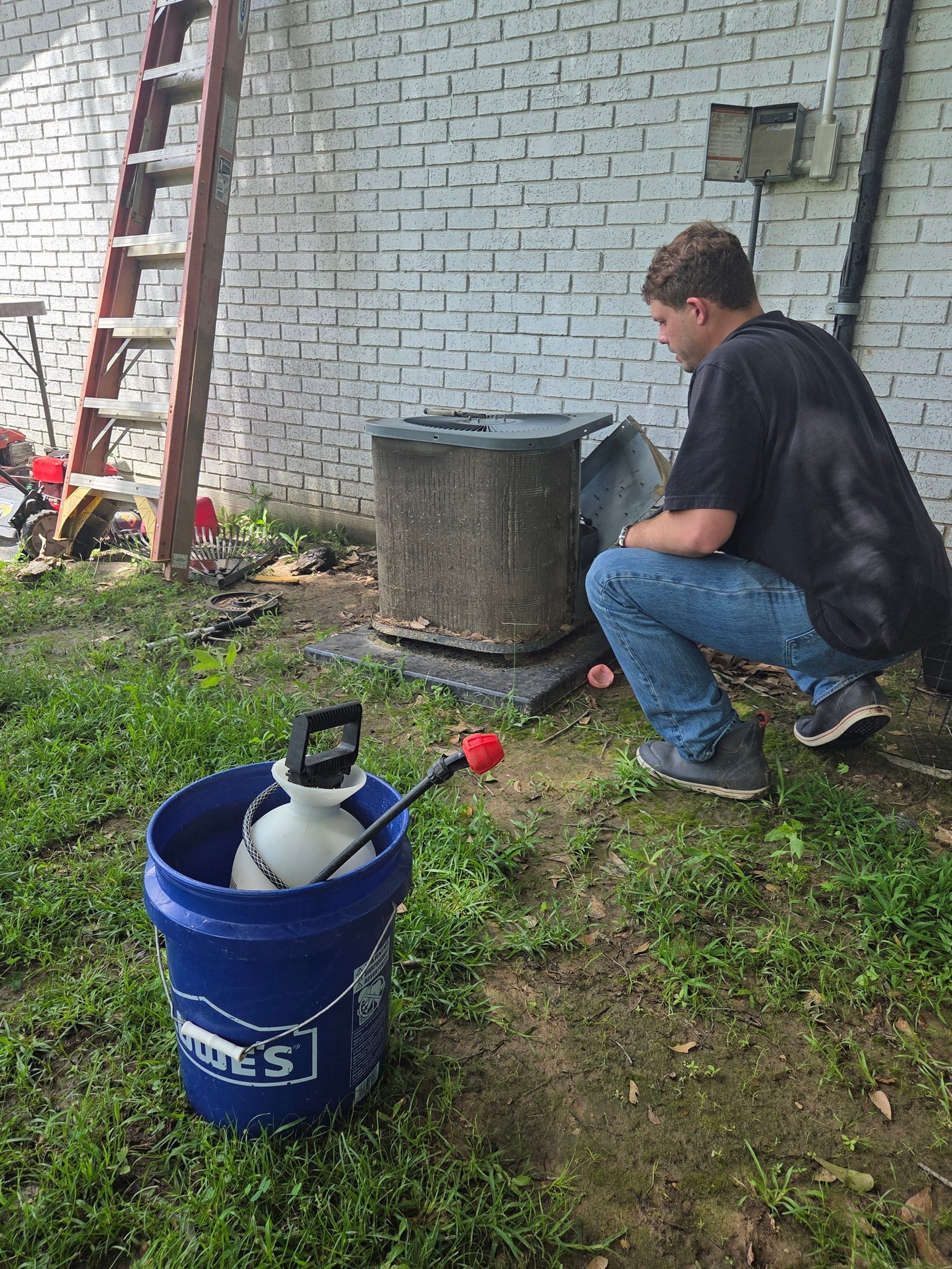 Person crouches by a brick wall next to a blue bucket and a ladder in a yard.