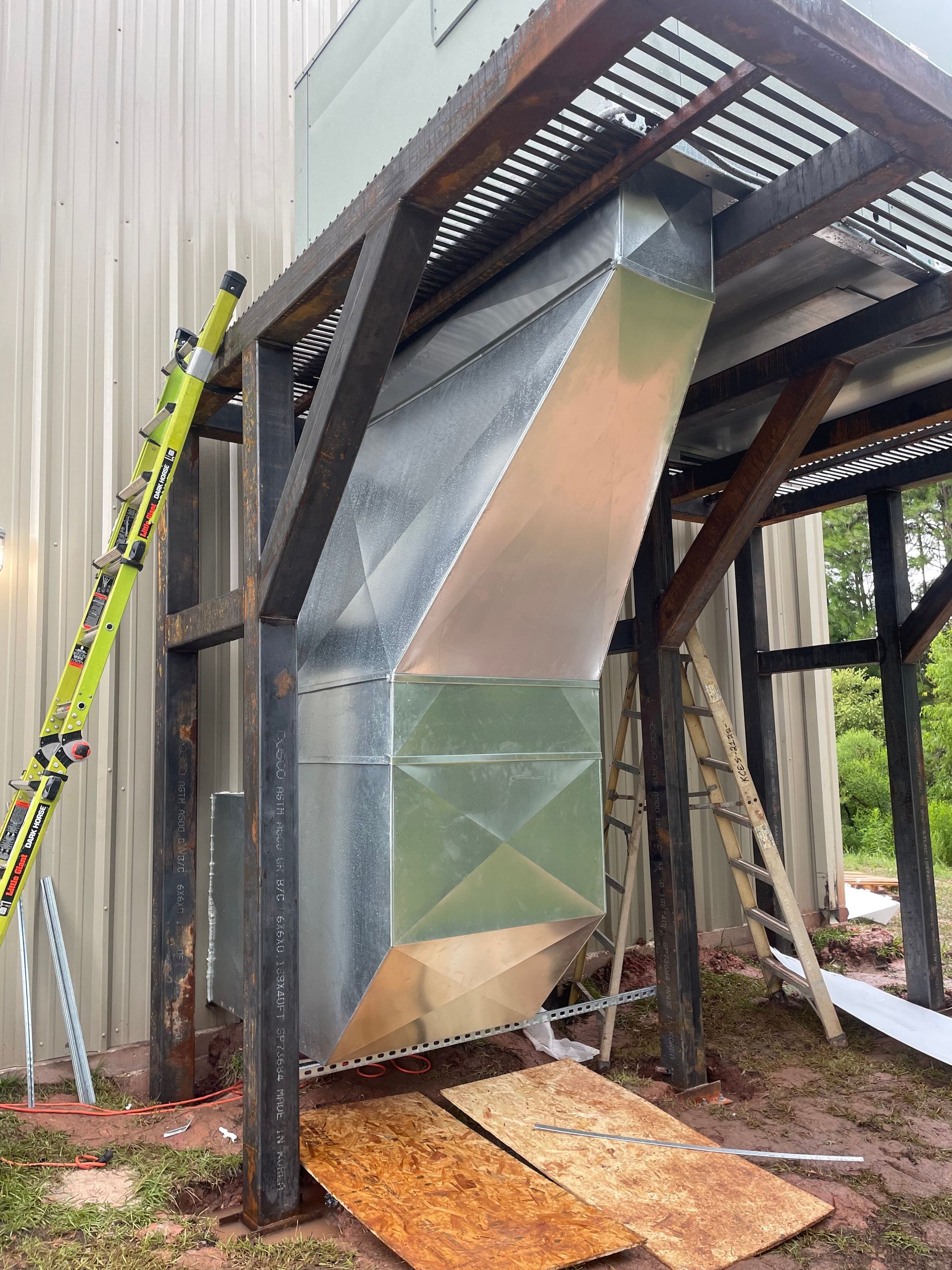 Metal ductwork under a raised platform beside a corrugated building, with a ladder leaning nearby.