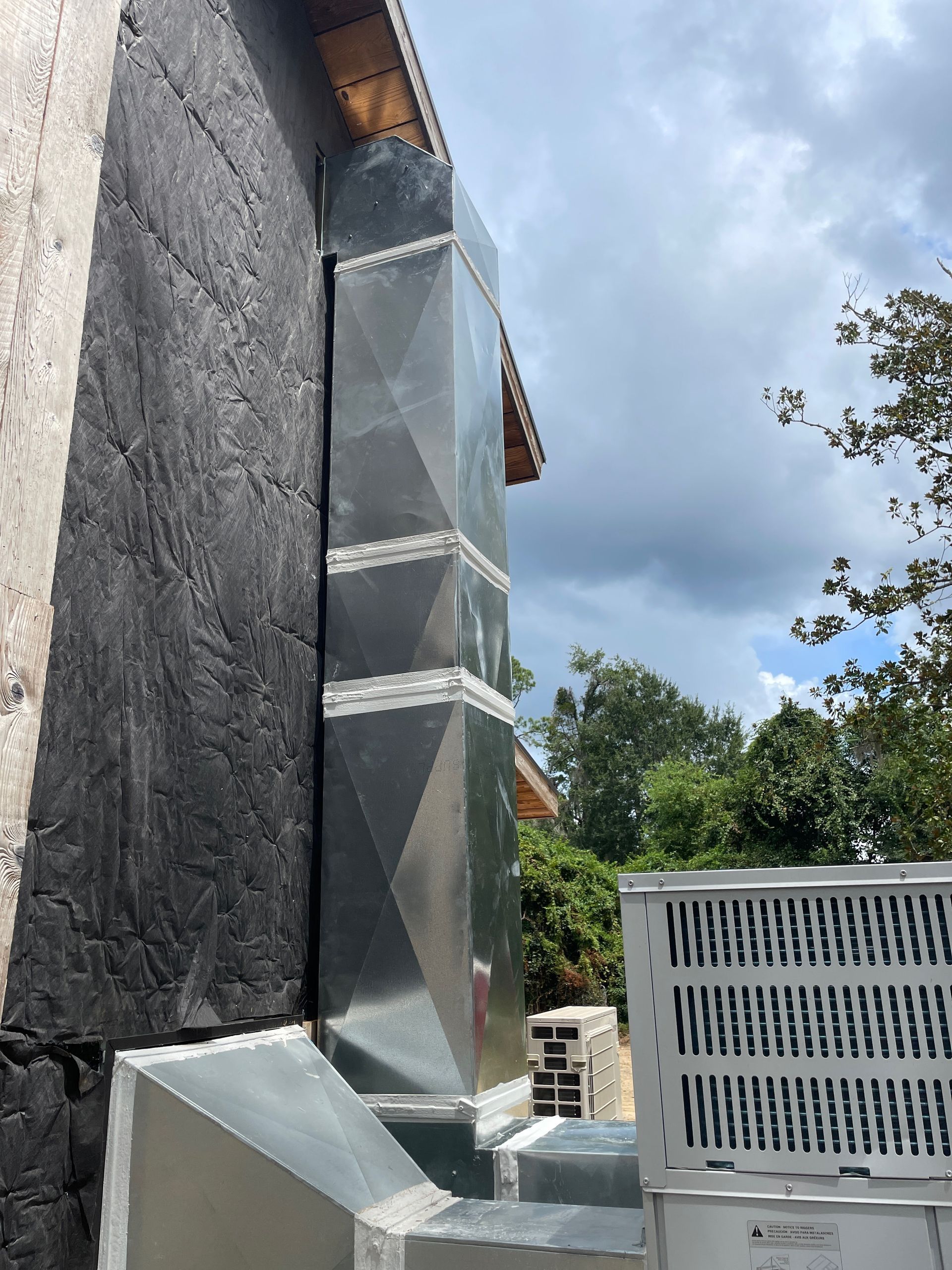 Metal HVAC duct beside a dark wall and rooftop unit against a cloudy sky