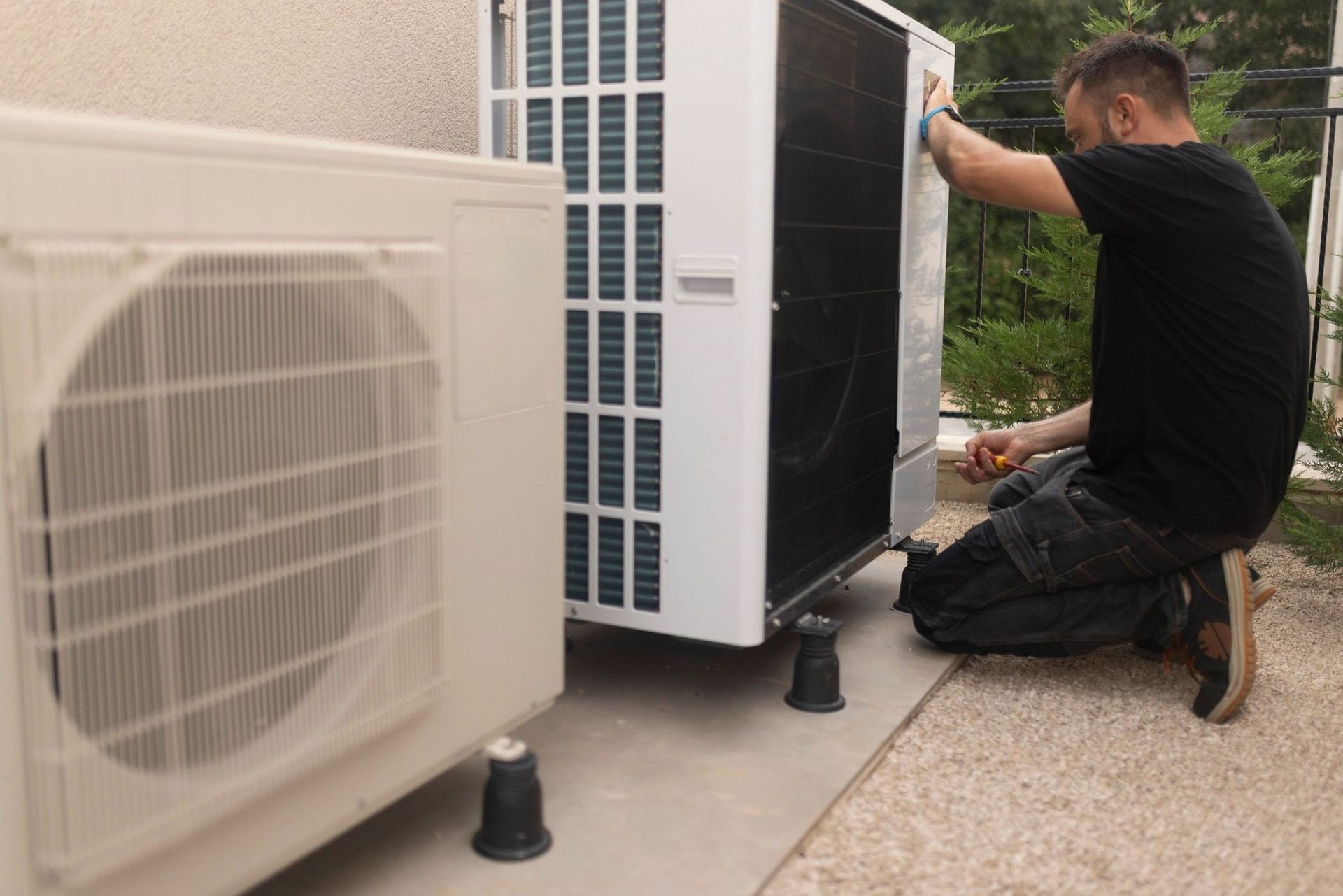 Technician kneeling beside a large outdoor HVAC unit, working on its side panel near a house.