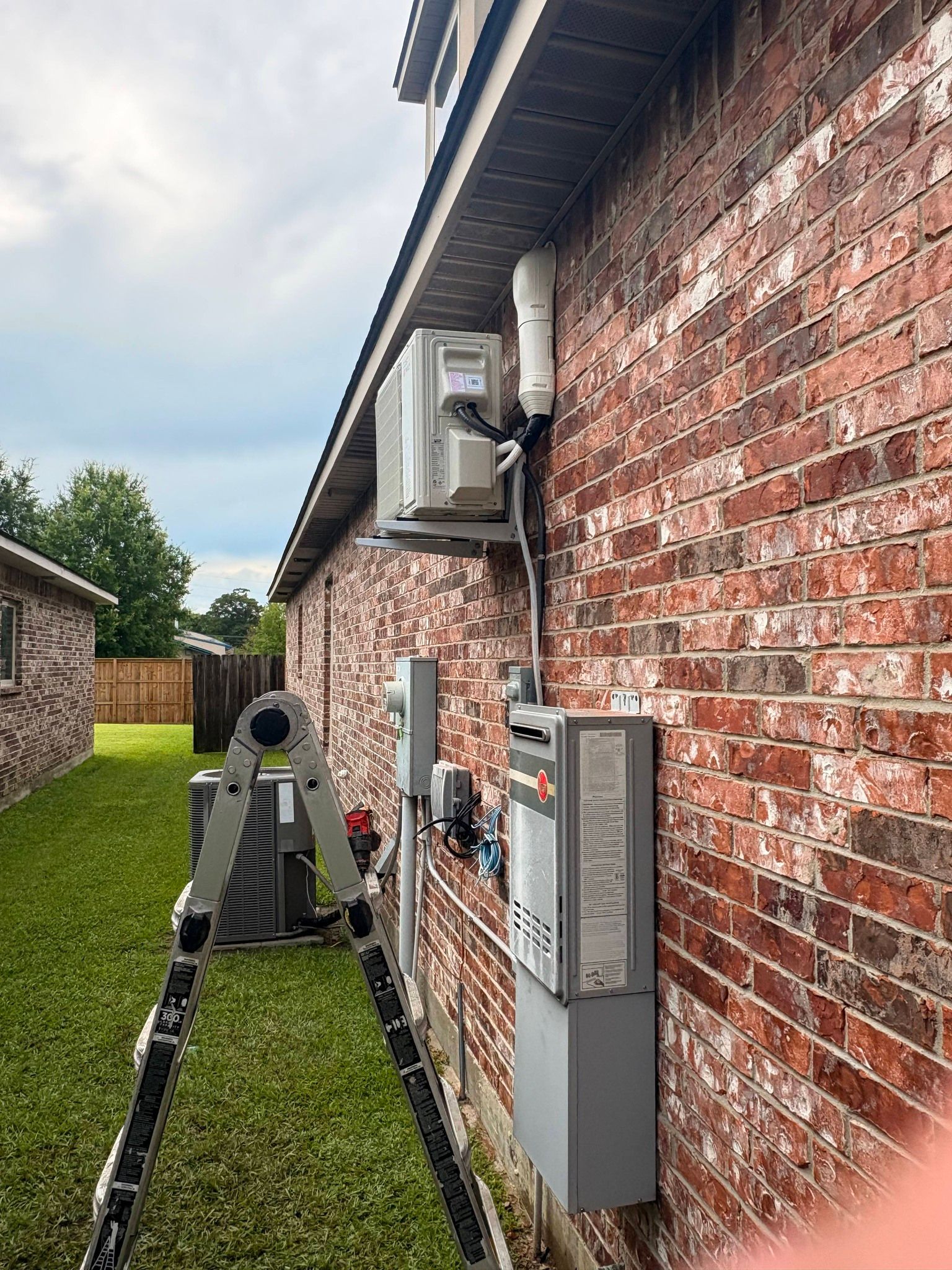 Brick house side wall with utility meters, wiring, and a person on a lawn mower beside the house