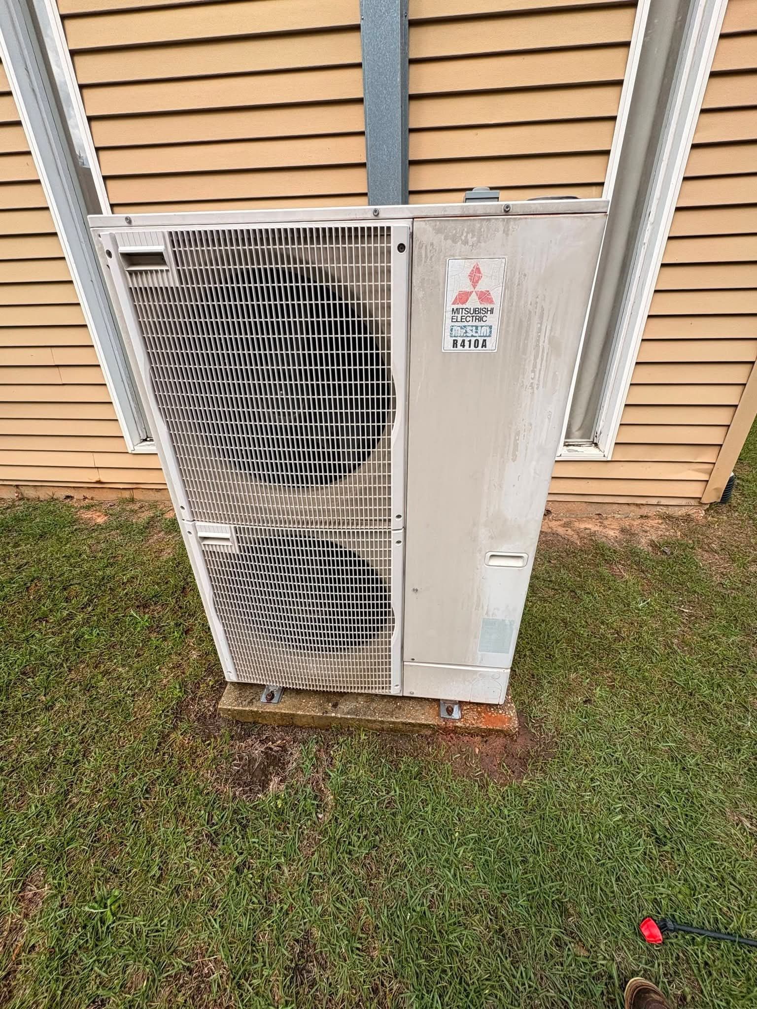 Outdoor HVAC unit leaning against a house wall on grass, with a bent metal grille and rusted base.