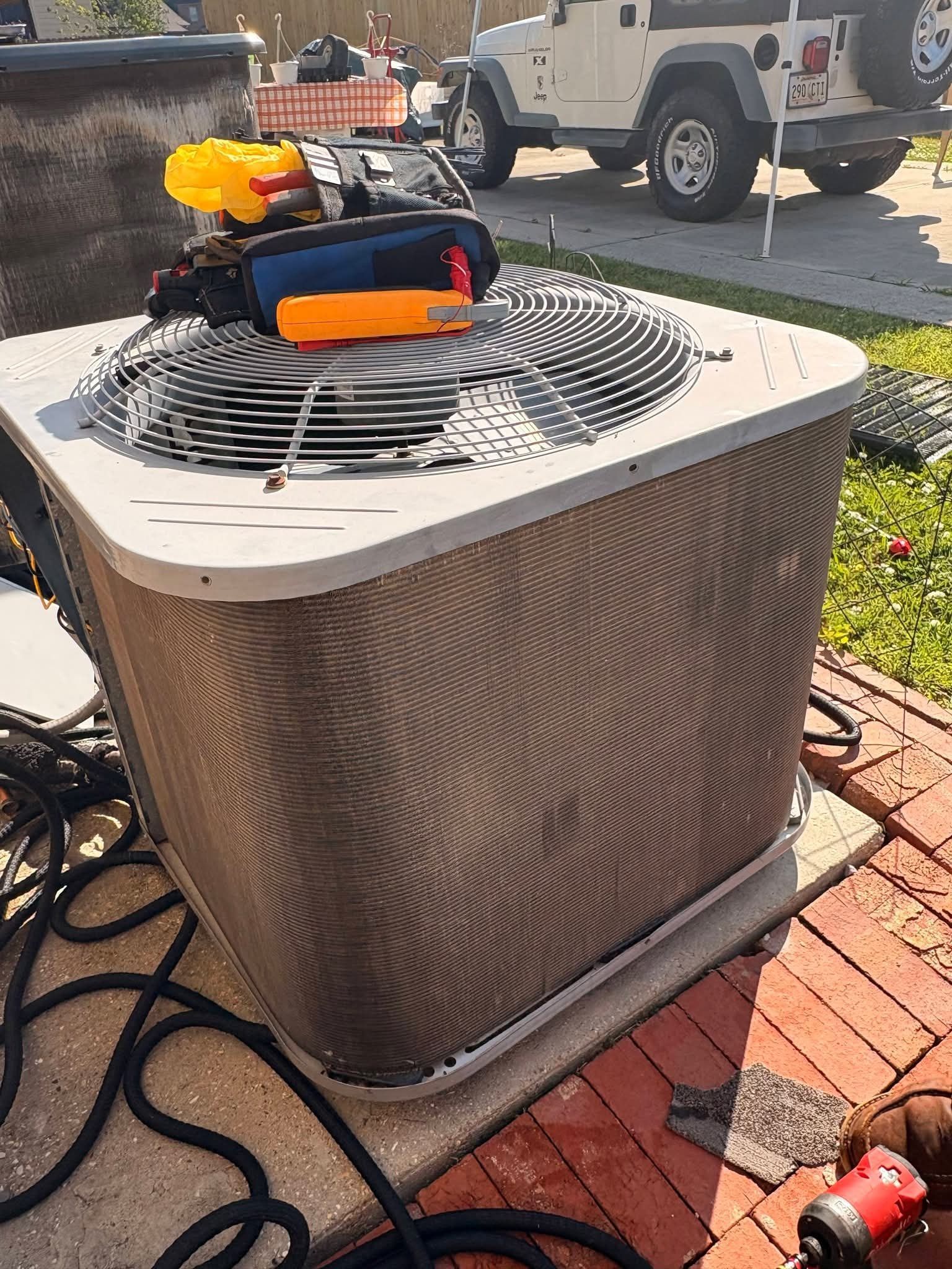 Outdoor HVAC condenser unit with a small orange-and-black toy car on top, near a brick walkway and hose.