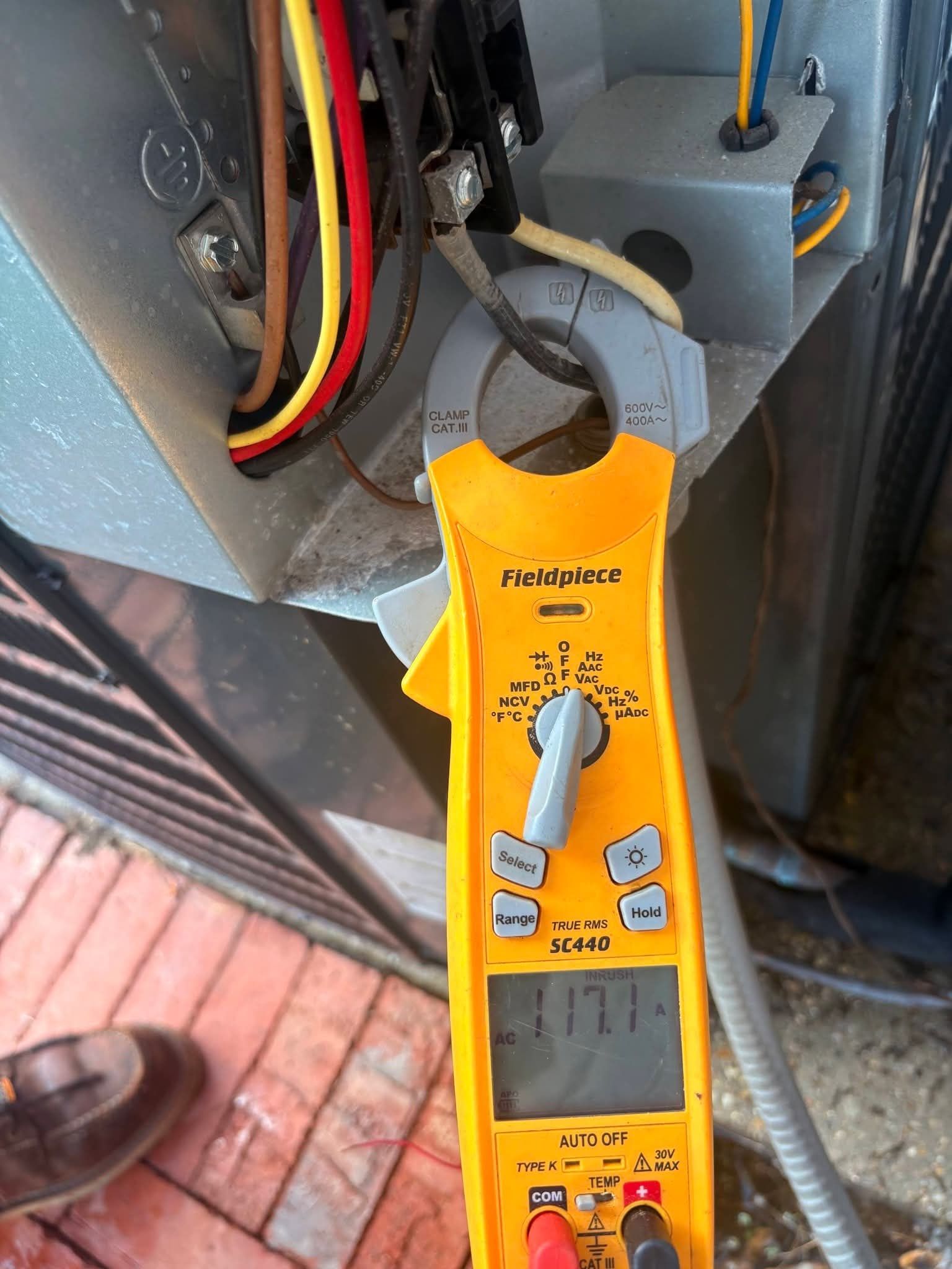 Yellow digital clamp meter measuring electrical wires beside a gray panel and brick floor.