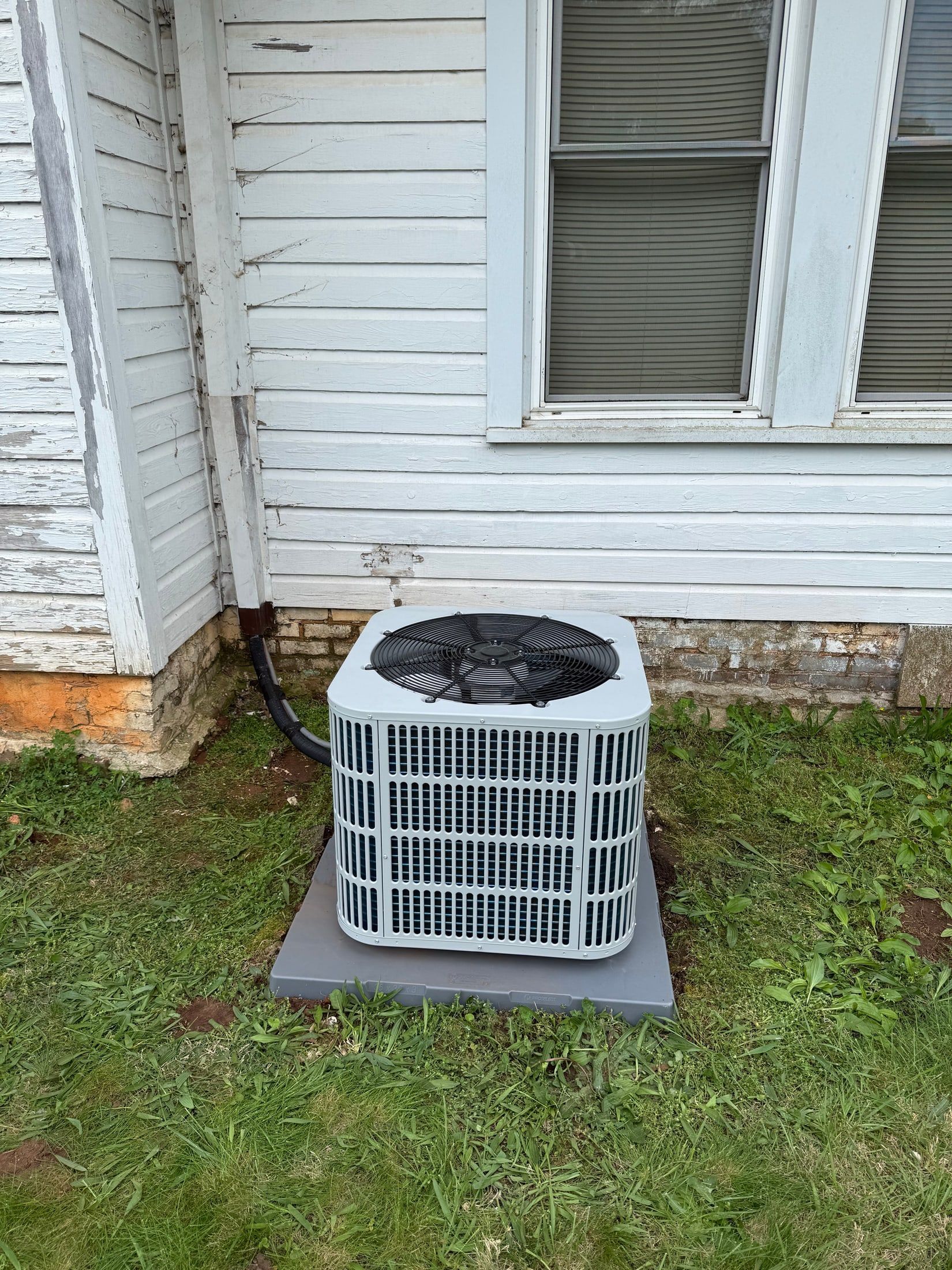 Outdoor air conditioner unit beside a white house with peeling siding and a small patch of grass