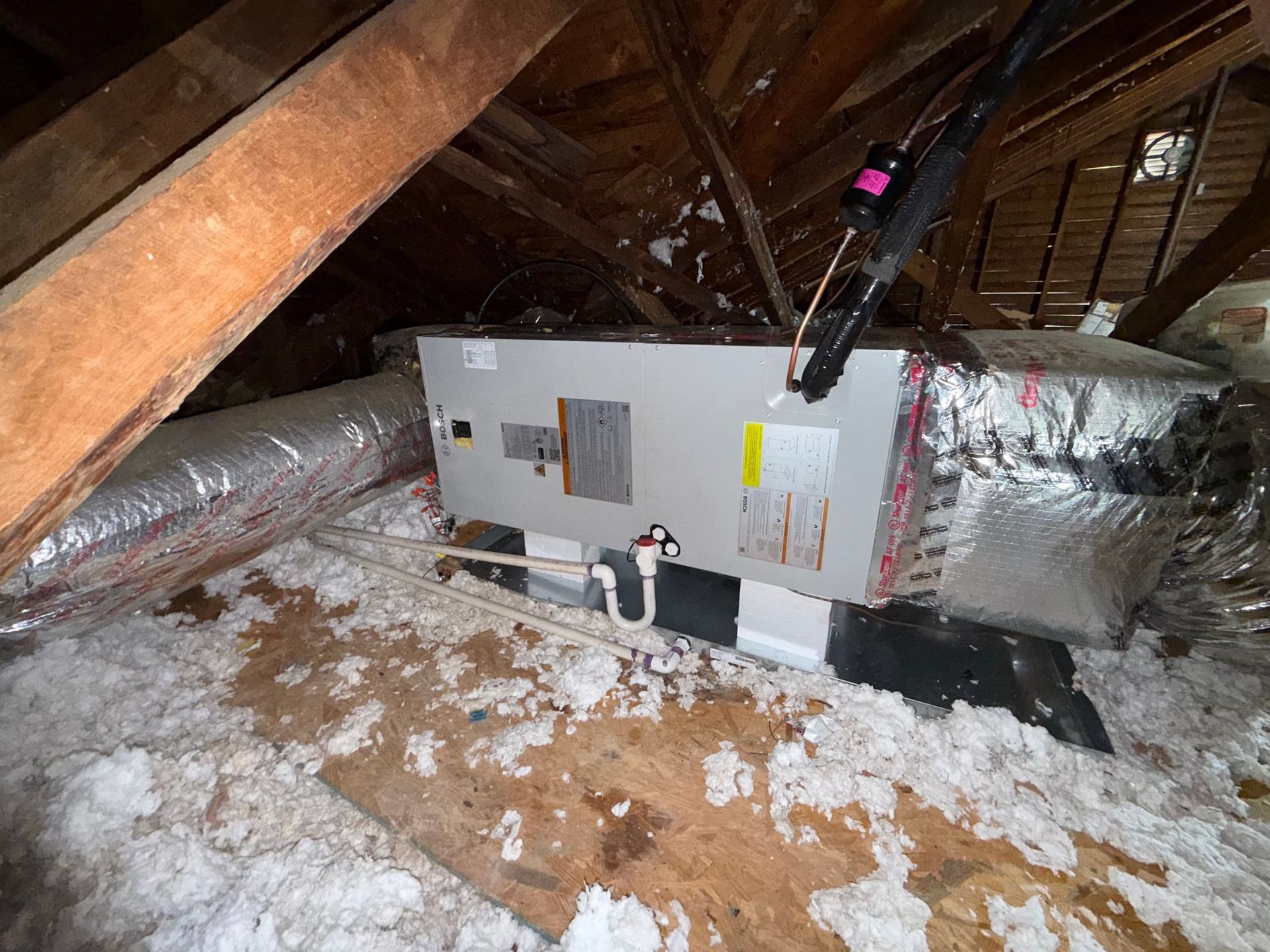 Attic HVAC unit wrapped in insulation, with exposed wooden rafters and snowy debris on the floor
