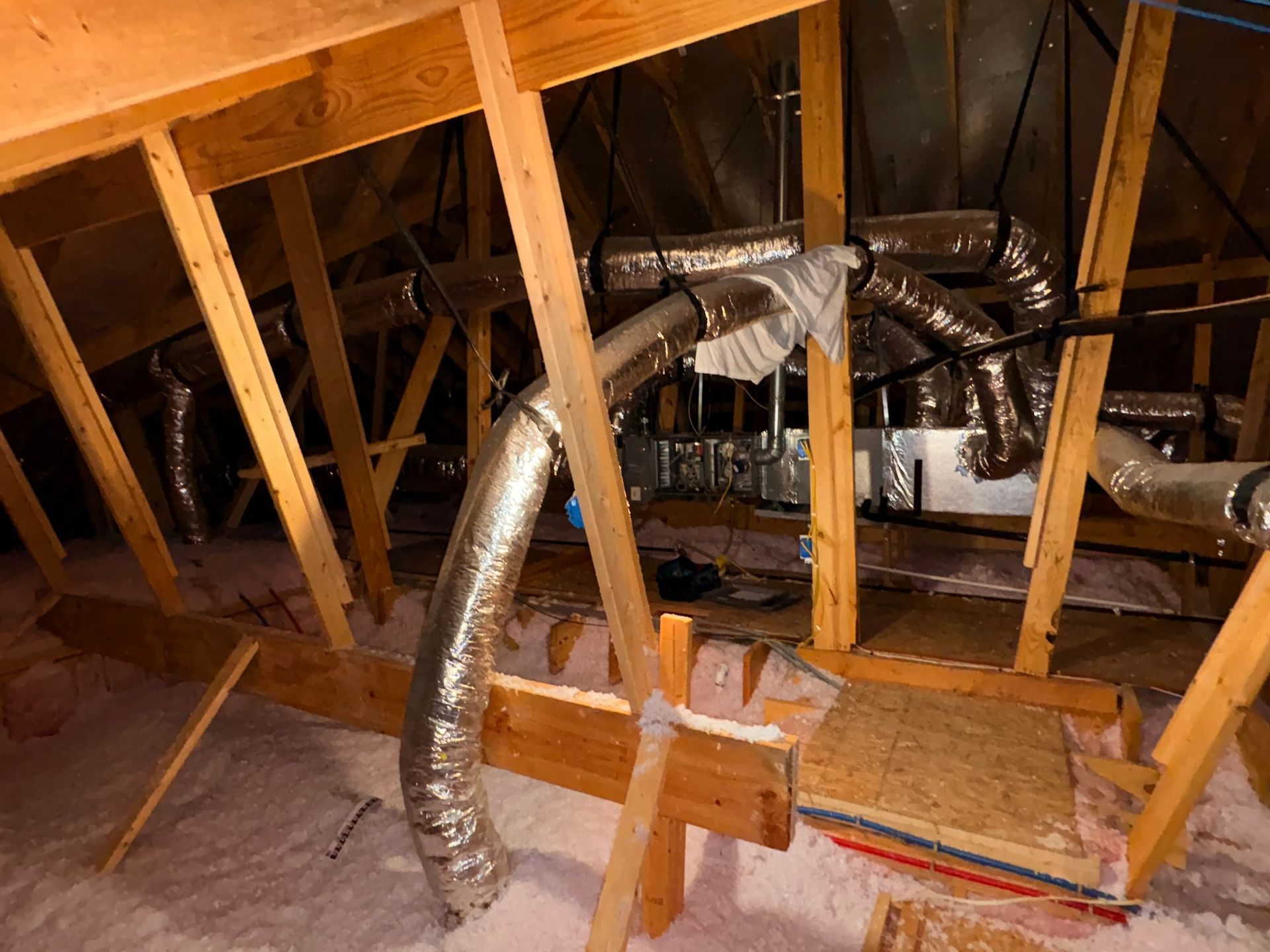 Attic with exposed wooden rafters and shiny HVAC ductwork running through the insulation.