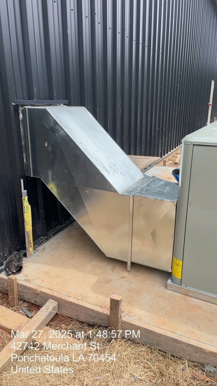 Metal HVAC duct with a sloped cover beside a corrugated black wall and a gray utility box outdoors