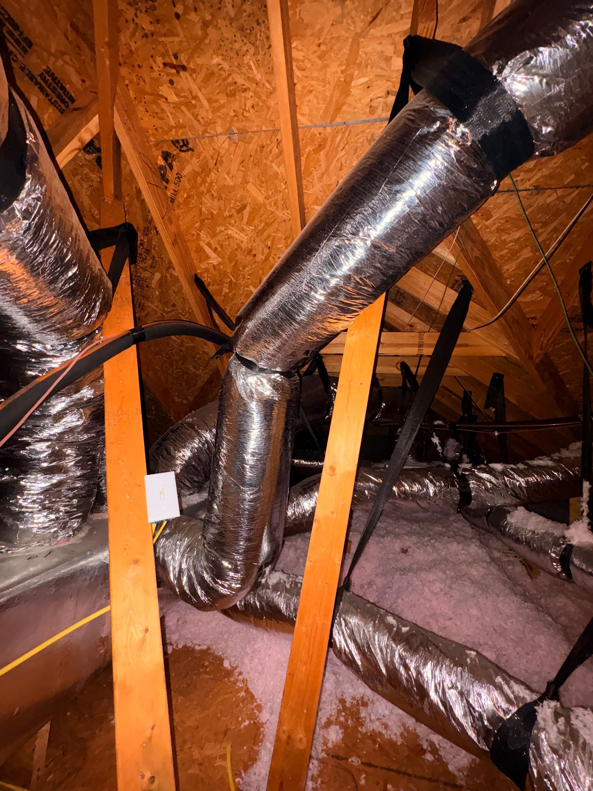 Insulated attic ductwork and vents beneath wooden roof rafters, with orange support straps and water stains