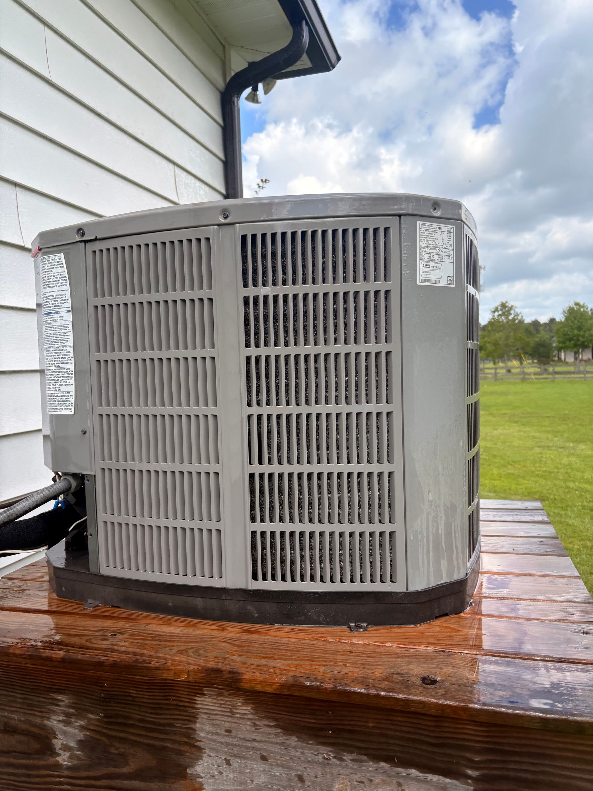 Outdoor air conditioner unit on a wooden platform beside a white house under cloudy sky