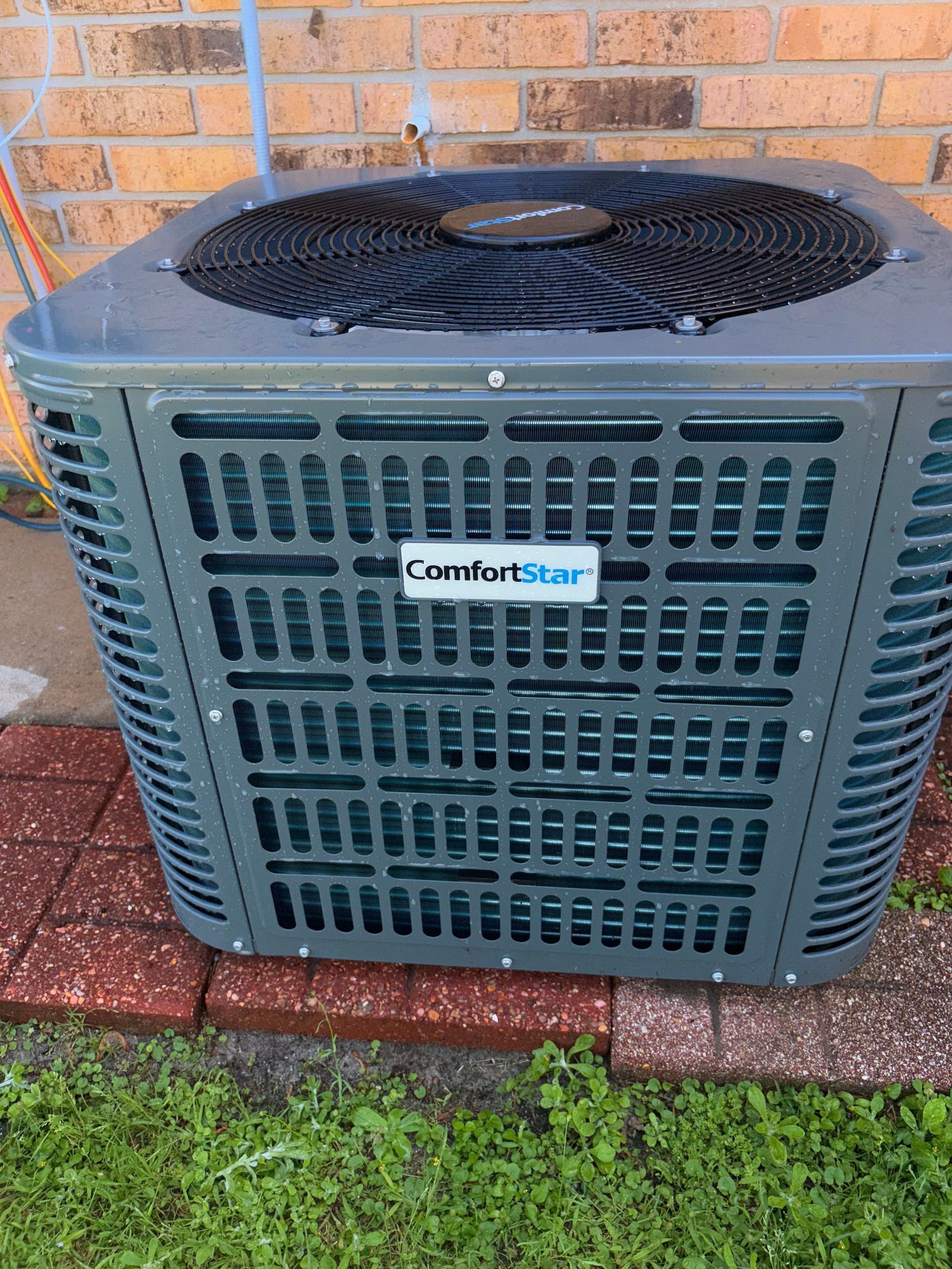Outdoor ComfortStar air conditioner unit beside a brick wall on a brick patio with grass nearby