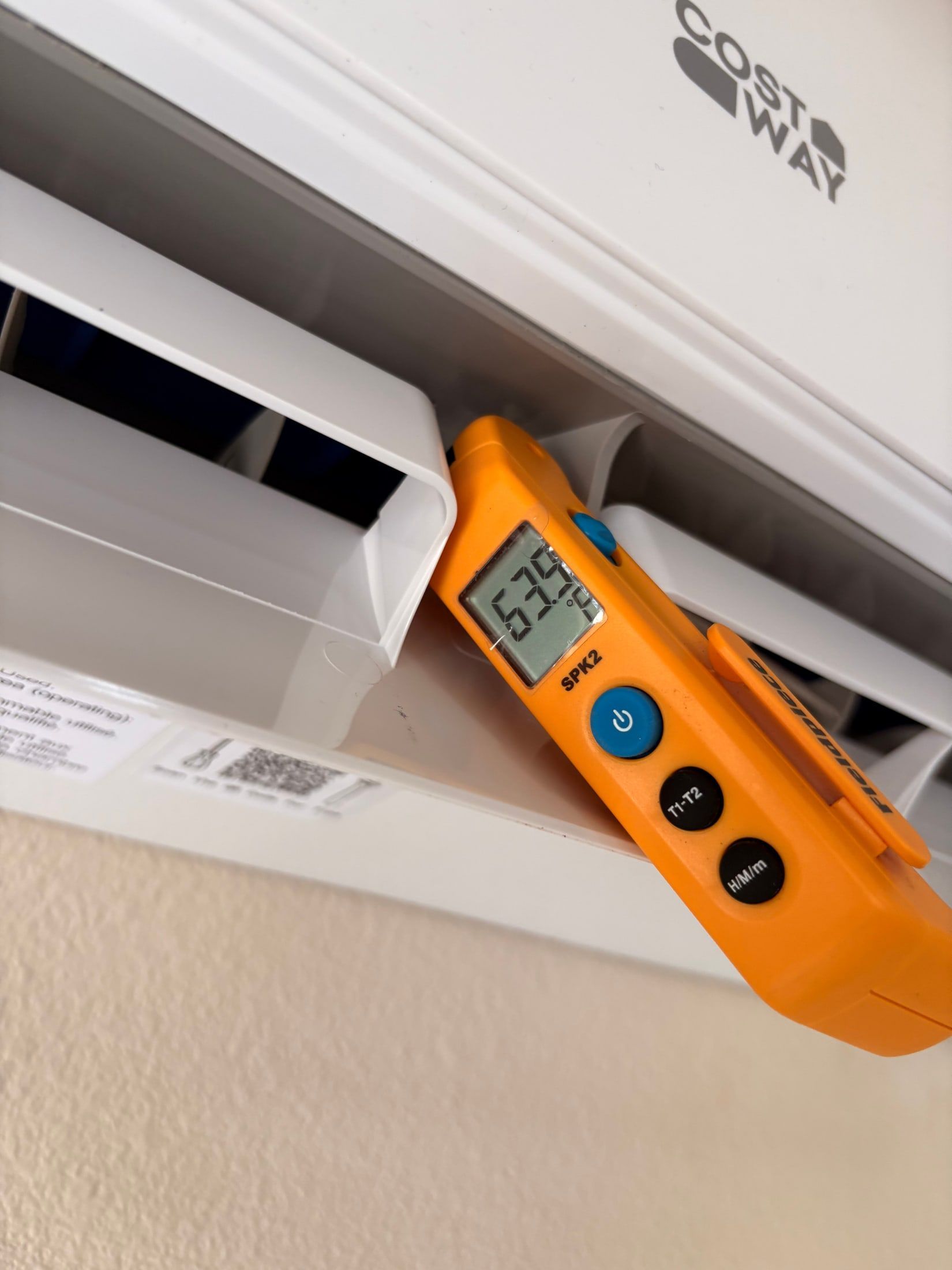Orange handheld meter measuring an air vent in a white ceiling vent opening
