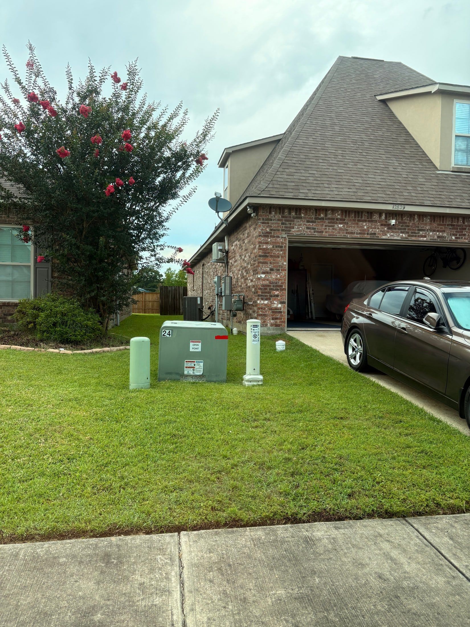 Suburban front yard with a utility box, flowering tree, driveway, and a black car beside a brick house.