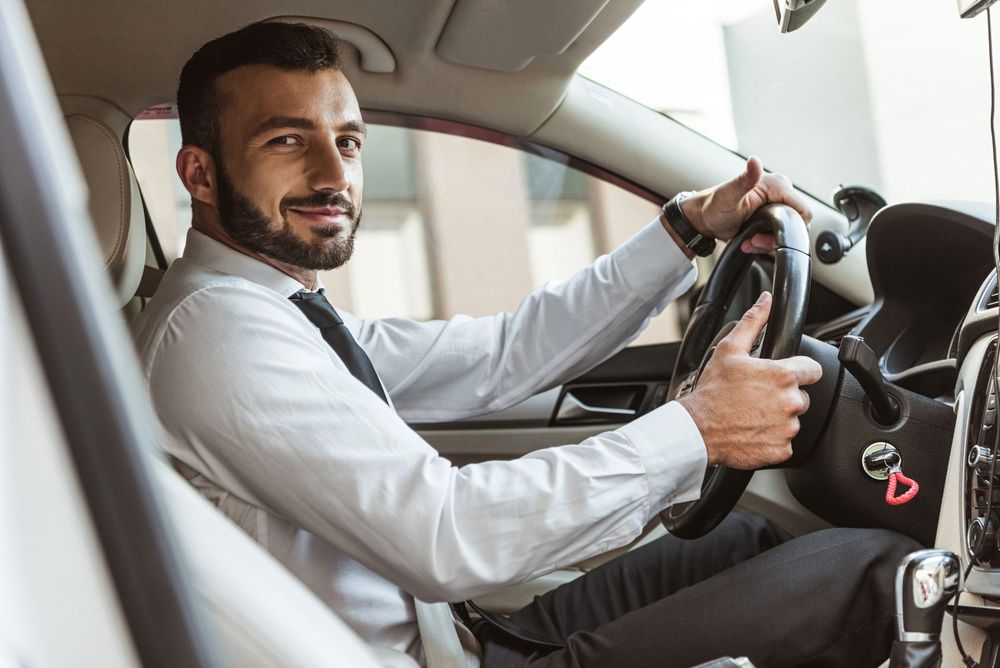 L'autista è seduto in macchina, sorridente, con il volante in una mano e la leva del cambio nell'altra.