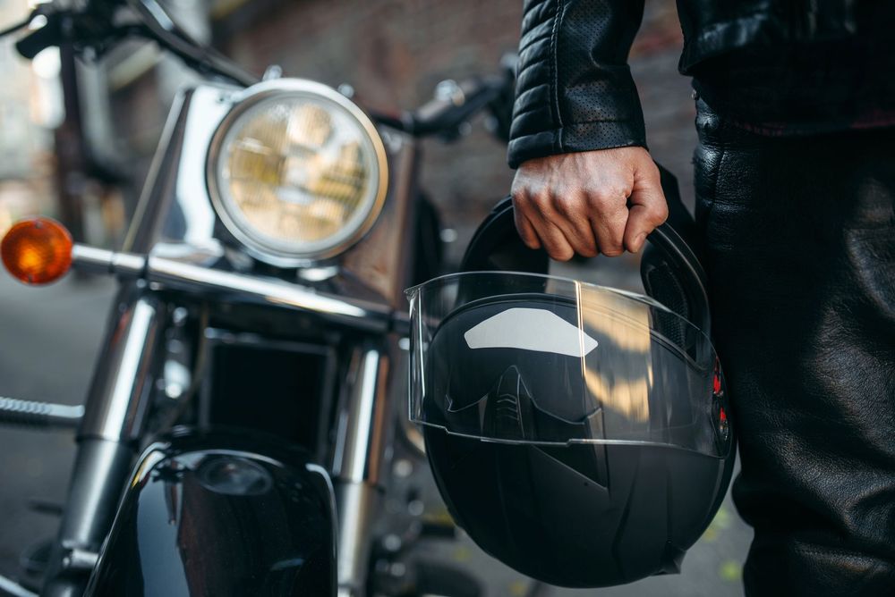 Persona che tiene in mano un casco nero accanto al faro di una motocicletta e alla manica di una giacca di pelle.