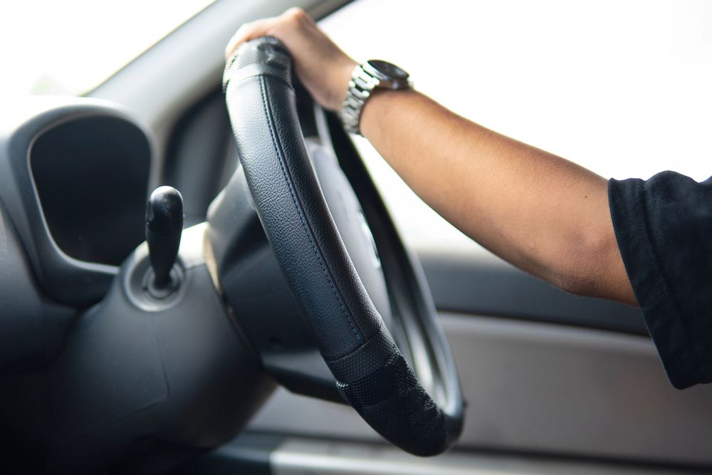 Mano sul volante all'interno di un'auto, con cruscotto e finestrino visibili.