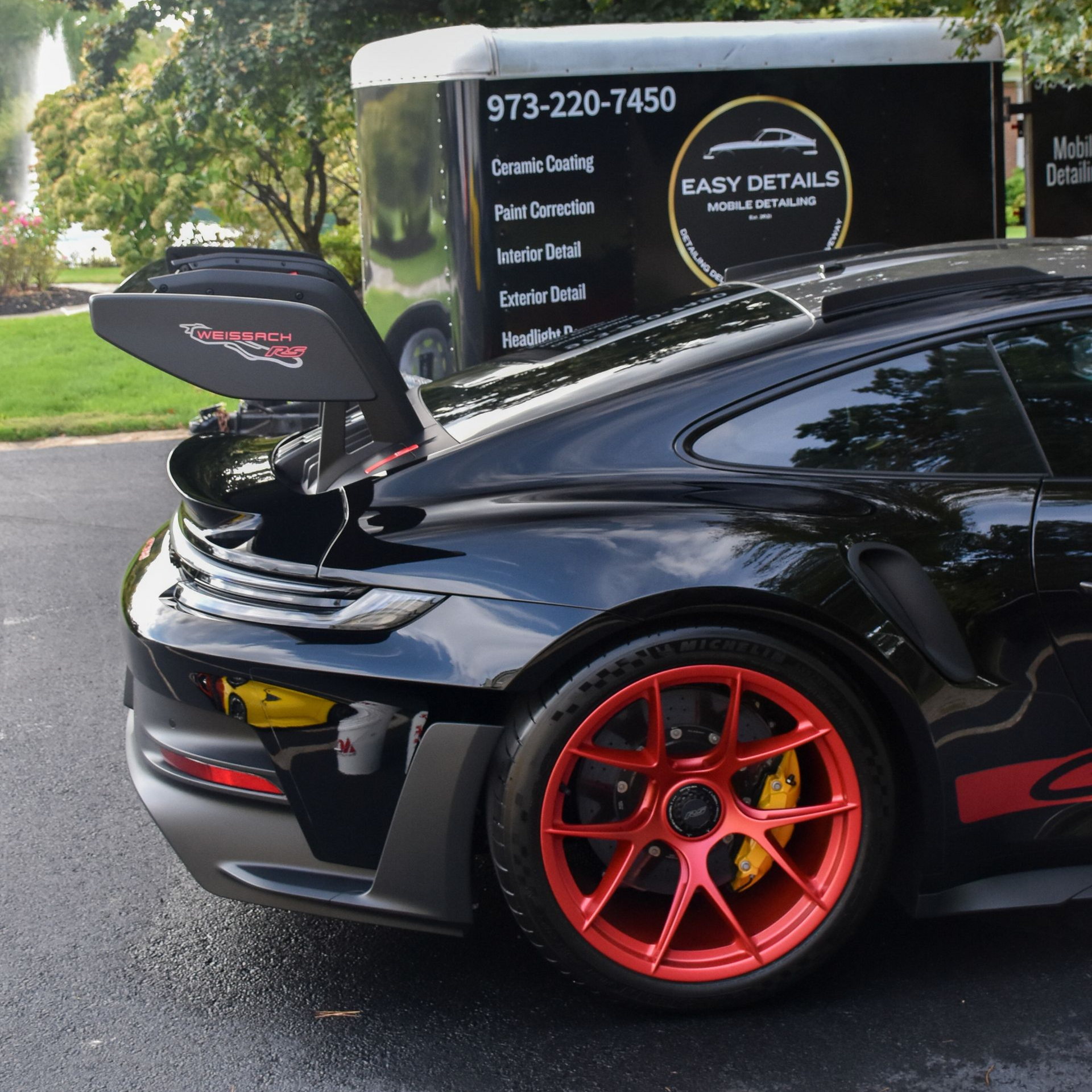 Black Porsche GT3 RS with red wheels, trunk open, parked next to a trailer in front of trees.