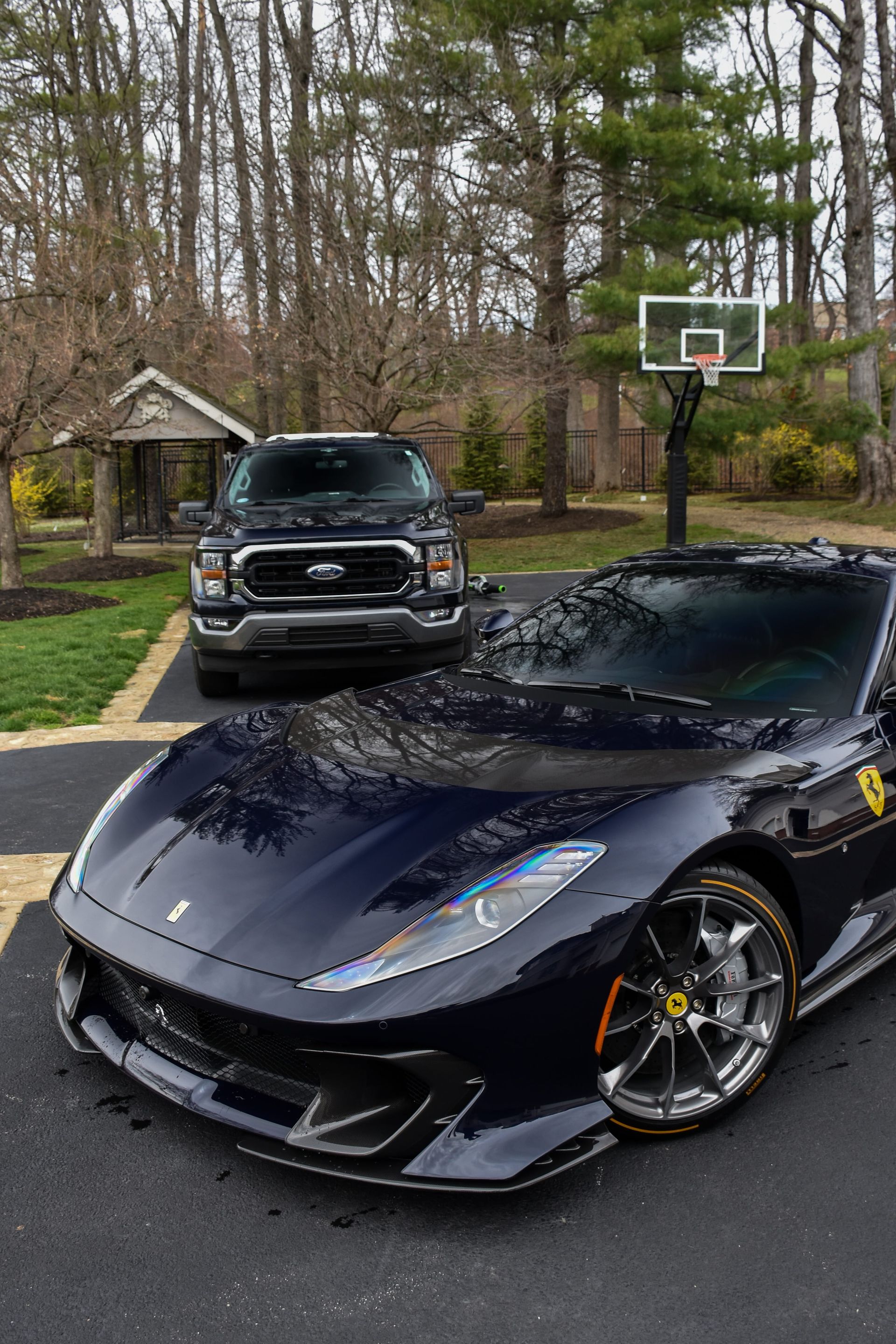 Black sports car parked in a driveway beside a pickup truck and basketball hoop in a wooded yard