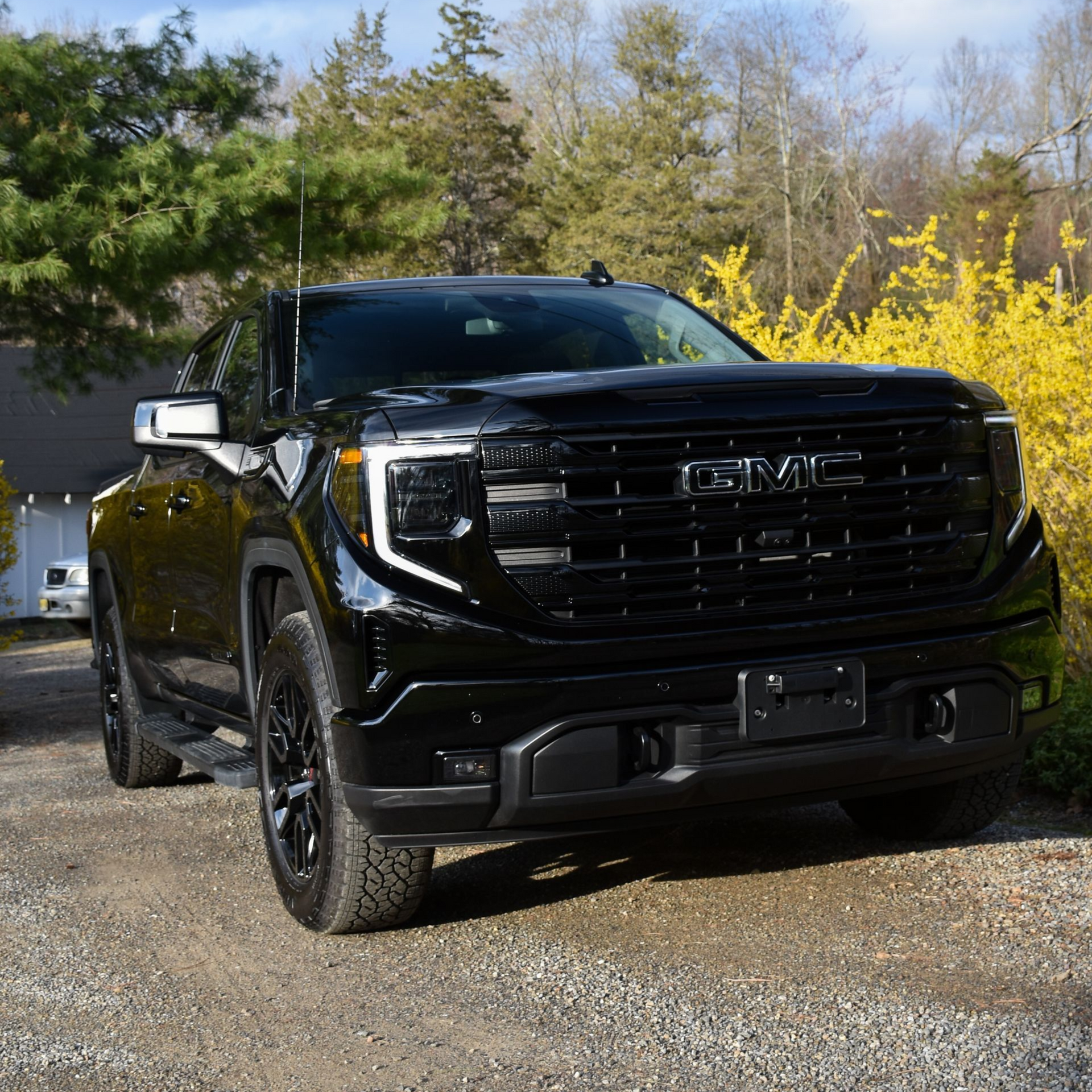Black pickup truck parked on a gravel driveway under a blue sky, with trees and yellow shrubs behind it