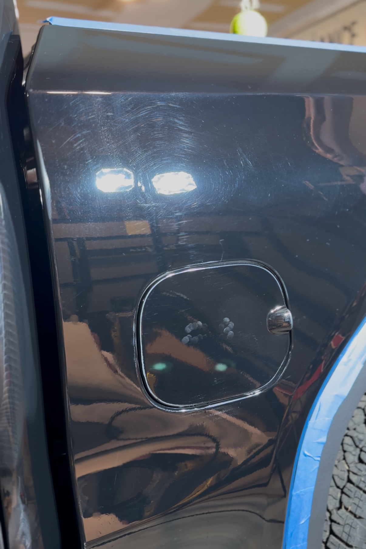 Close-up of a black car’s fuel door and rear wheel arch with blue painter’s tape along the edge