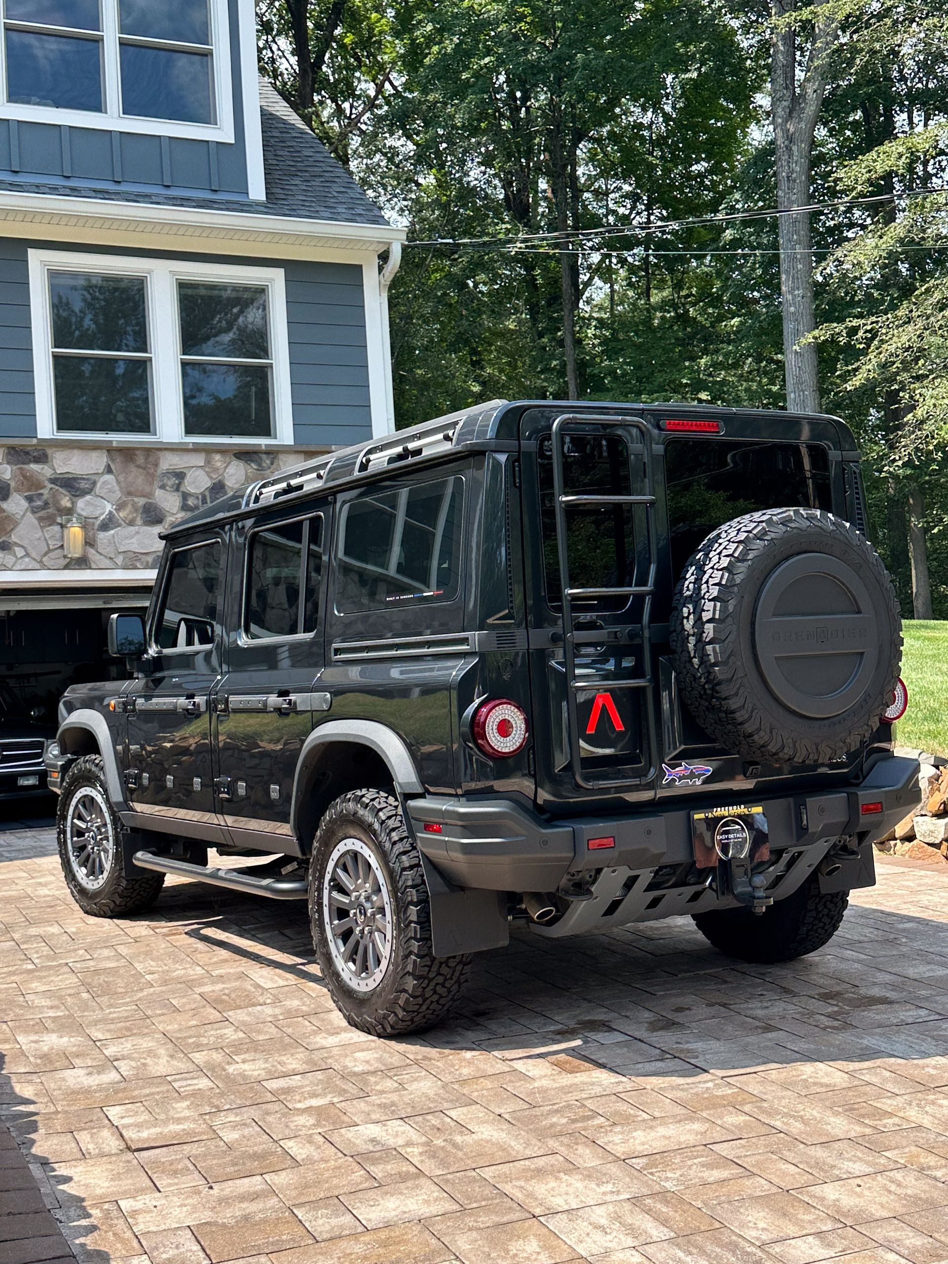 Black SUV with spare tire, ladder, parked on brick driveway in front of a house.