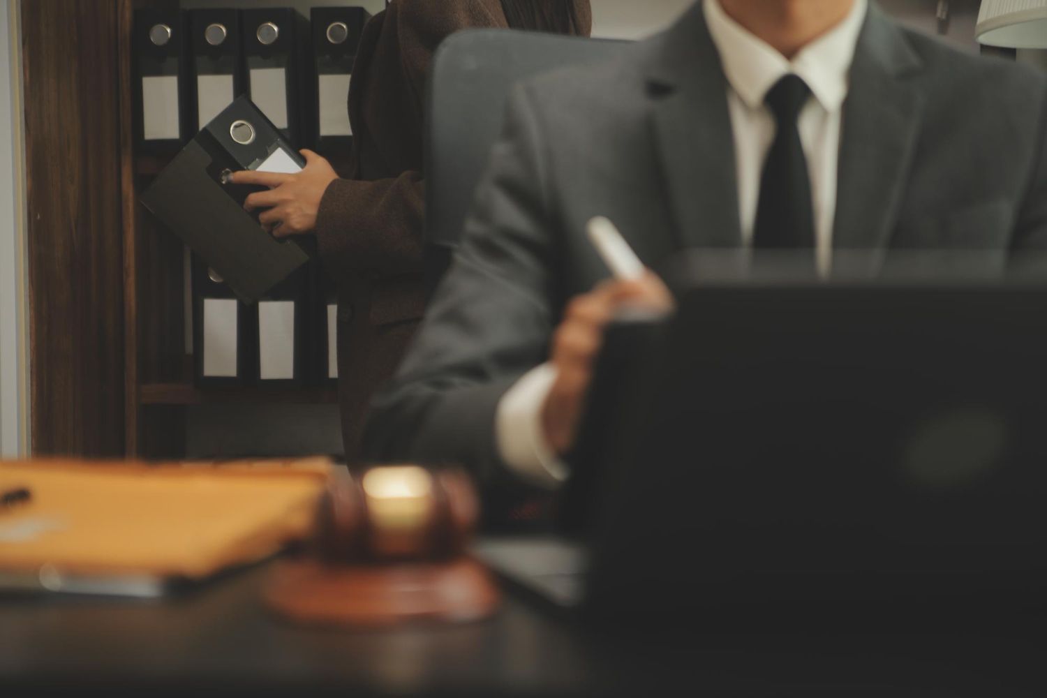 Office meeting scene with a suited person writing at a desk and another person entering near binders.