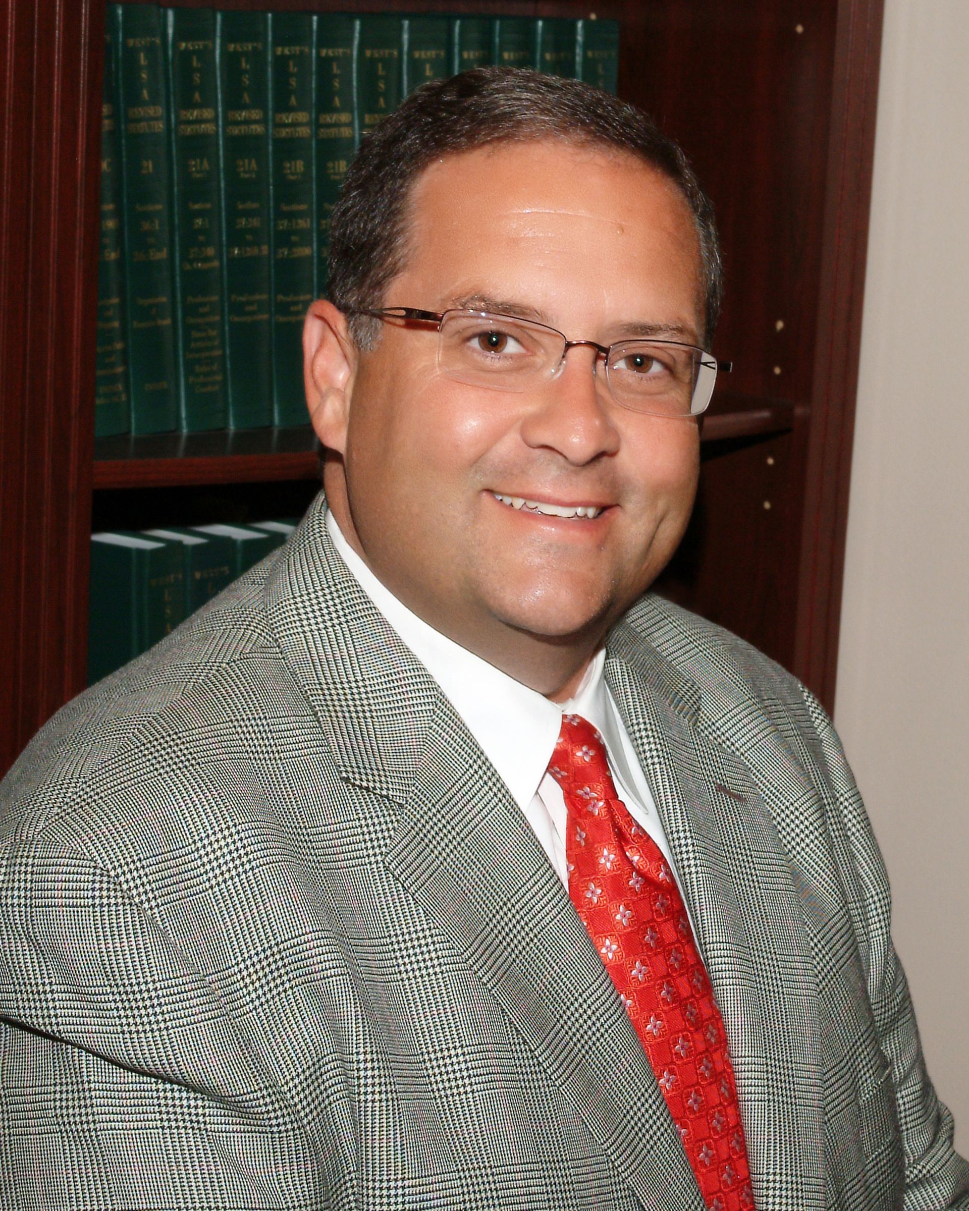 Man in glasses wearing a gray suit and red tie, seated in an office with bookshelves behind him