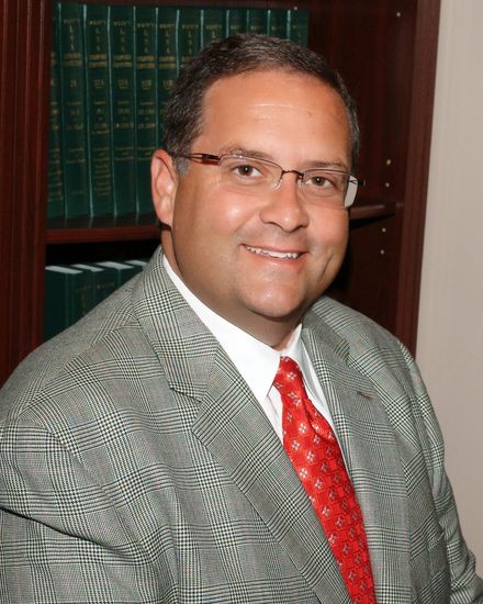 Man in glasses wearing a gray suit and red tie, seated in an office with bookshelves behind him