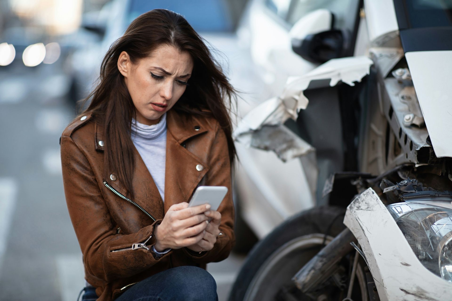 Woman in brown jacket checking her phone beside a damaged car after a collision