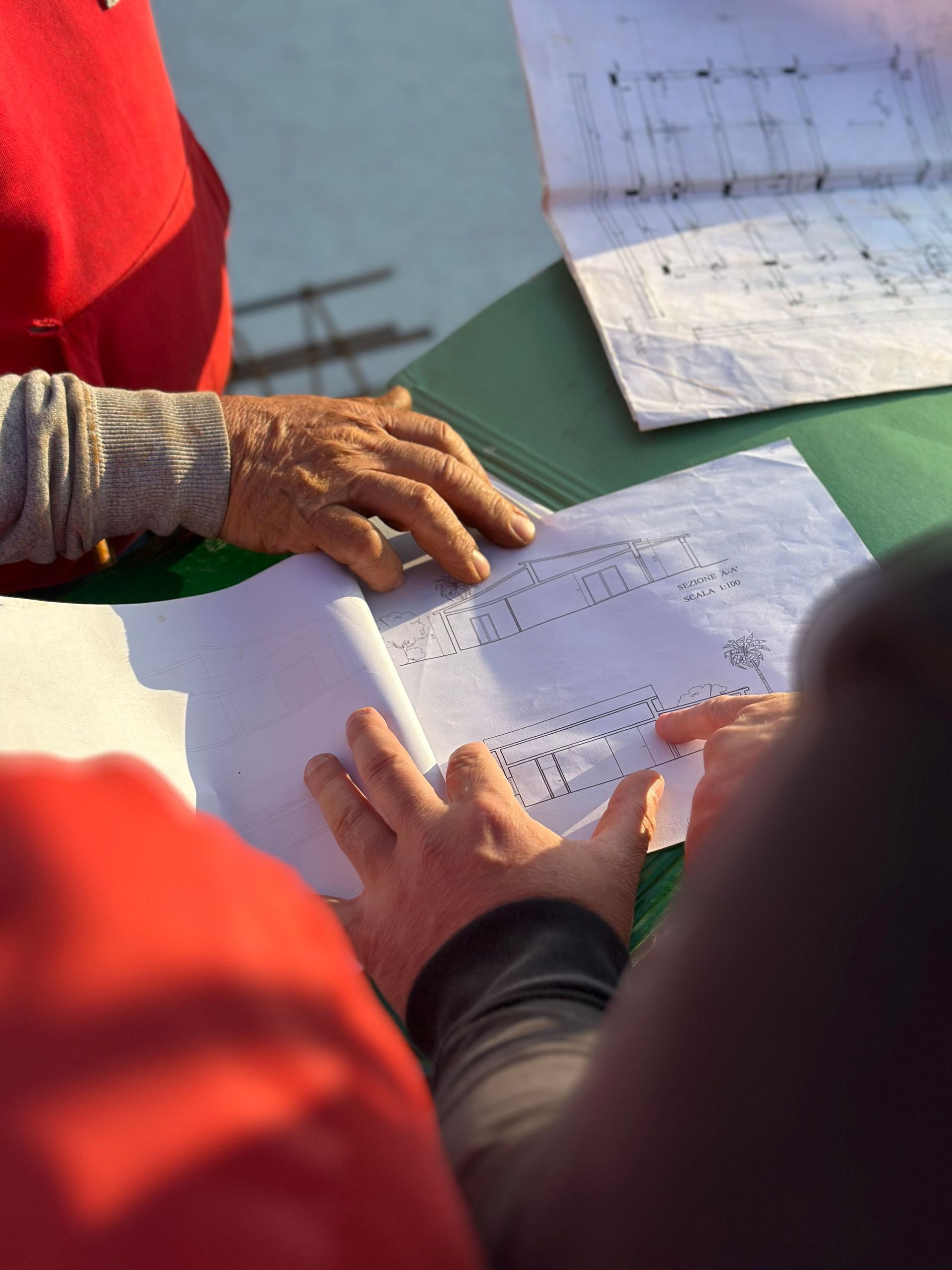 Hands and papers on a green table outdoors, with a red jacket in the foreground.