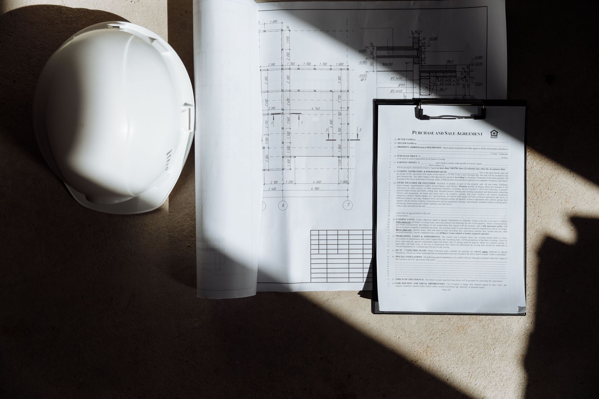 White hard hat beside architectural plans and a clipboard in sunlight on a desk