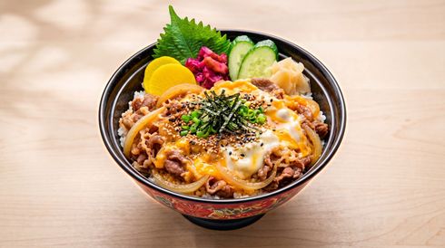 Bowl of beef and rice topped with melted cheese, herbs, and colorful pickles on a wooden table
