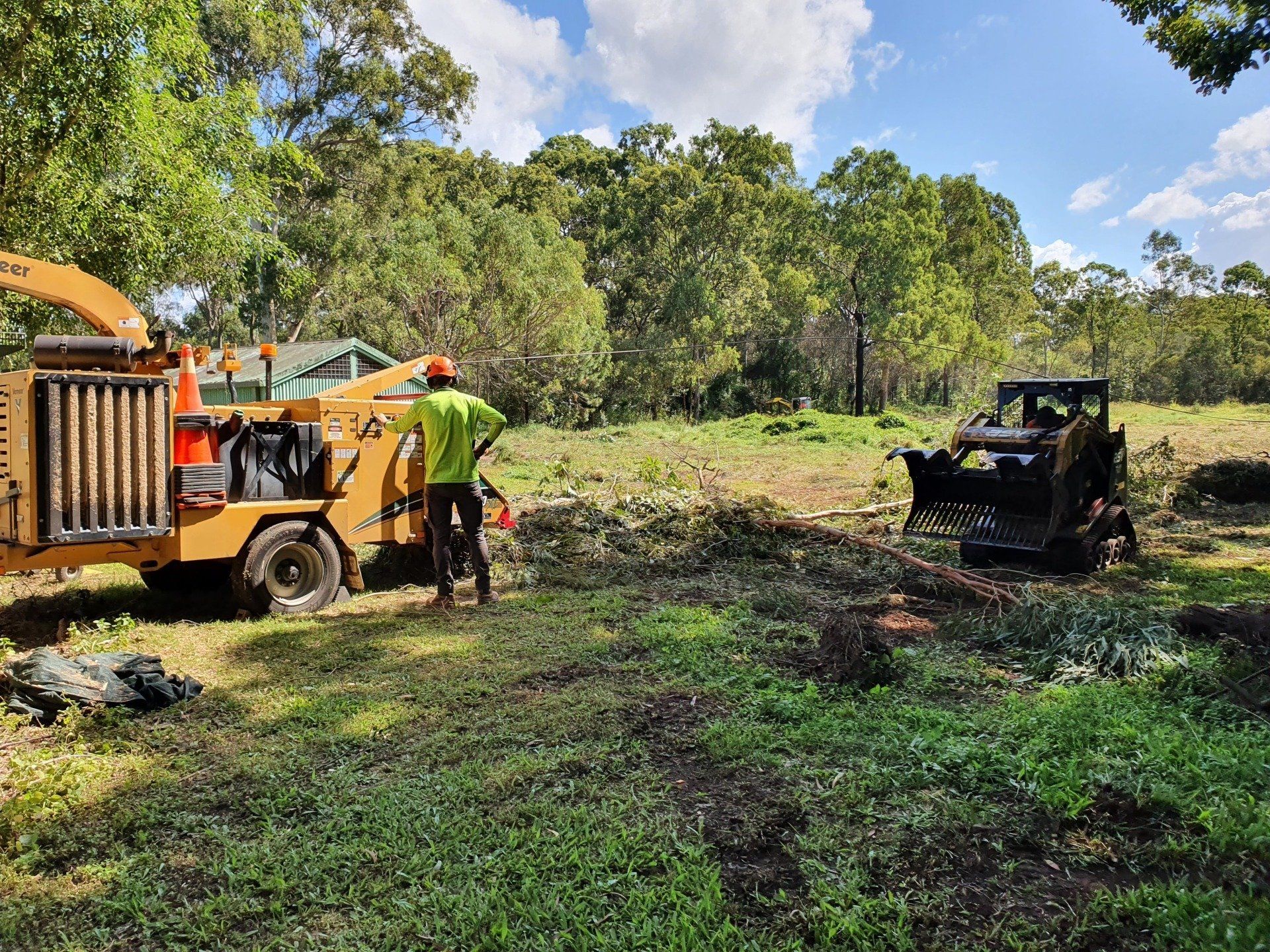 Workers beside an orange wood chipper and a black utility vehicle in a grassy clearing with trees