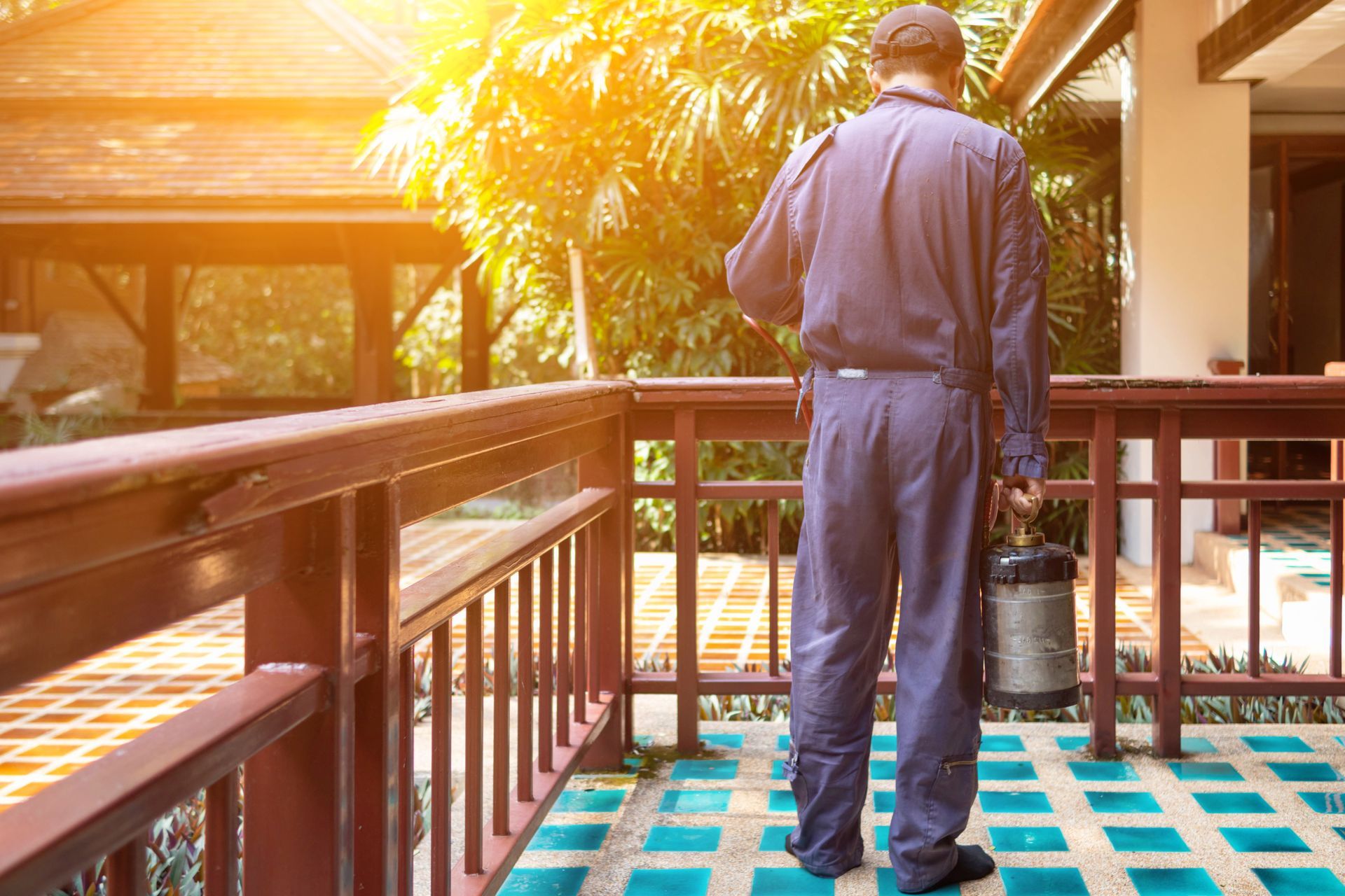 Technician spraying treatment around residential property exterior. Technician in blue uniform holding a sprayer performs pest control on a wooden deck near greenery.