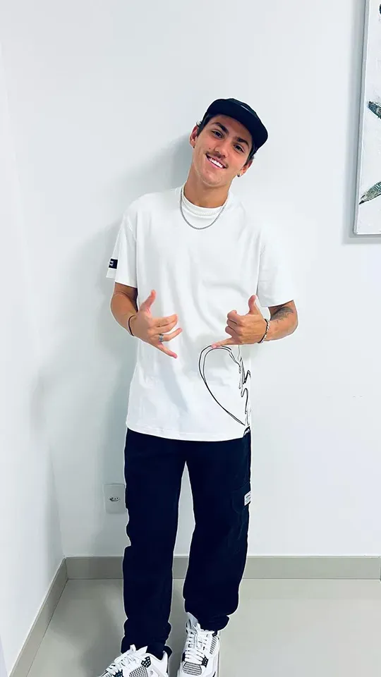 A young man wearing a white t-shirt and black pants is standing in front of a white wall.