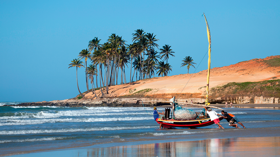 Una playa con pescadores empujando una barca A man is pulling a boat on a beach with palm trees in the background.