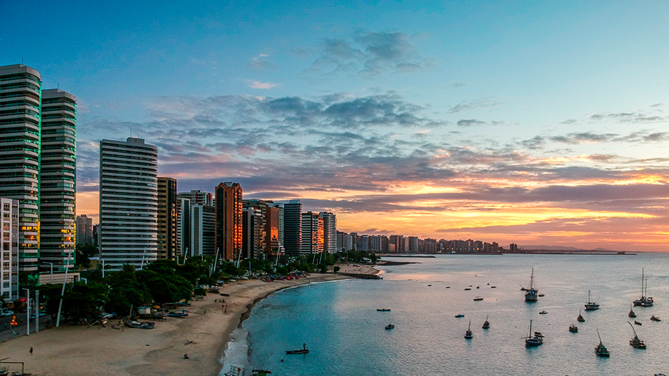Edificios alineados frente a la costa de Ceará An aerial view of a city and a beach at sunset with boats in the water.