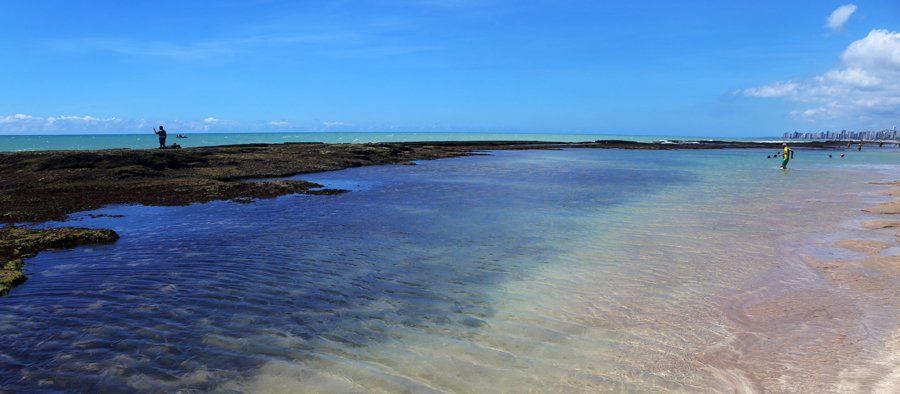 Le récif corallien dessine des piscines naturelles le long de la côte, et on aperçoit des pêcheurs au loin.