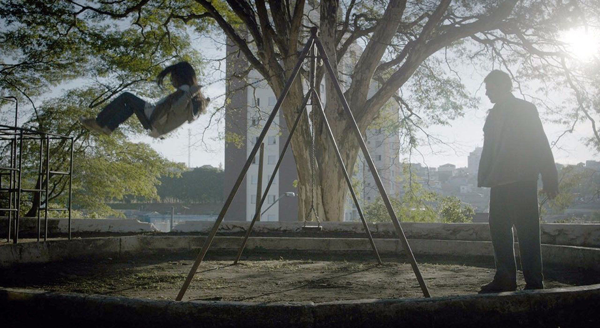 Ein Standbild aus dem Film „A sombra do pai“, gedreht in Butantã A man is standing next to a swing set with a child on it.