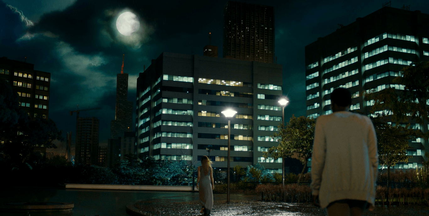 Eine Frau geht im Regen auf einem Platz in Alphaville spazieren A man and a woman are walking in the rain in front of a city at night.