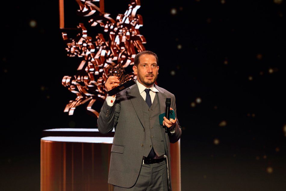 A man in a suit and tie is holding a microphone and a trophy on a stage.