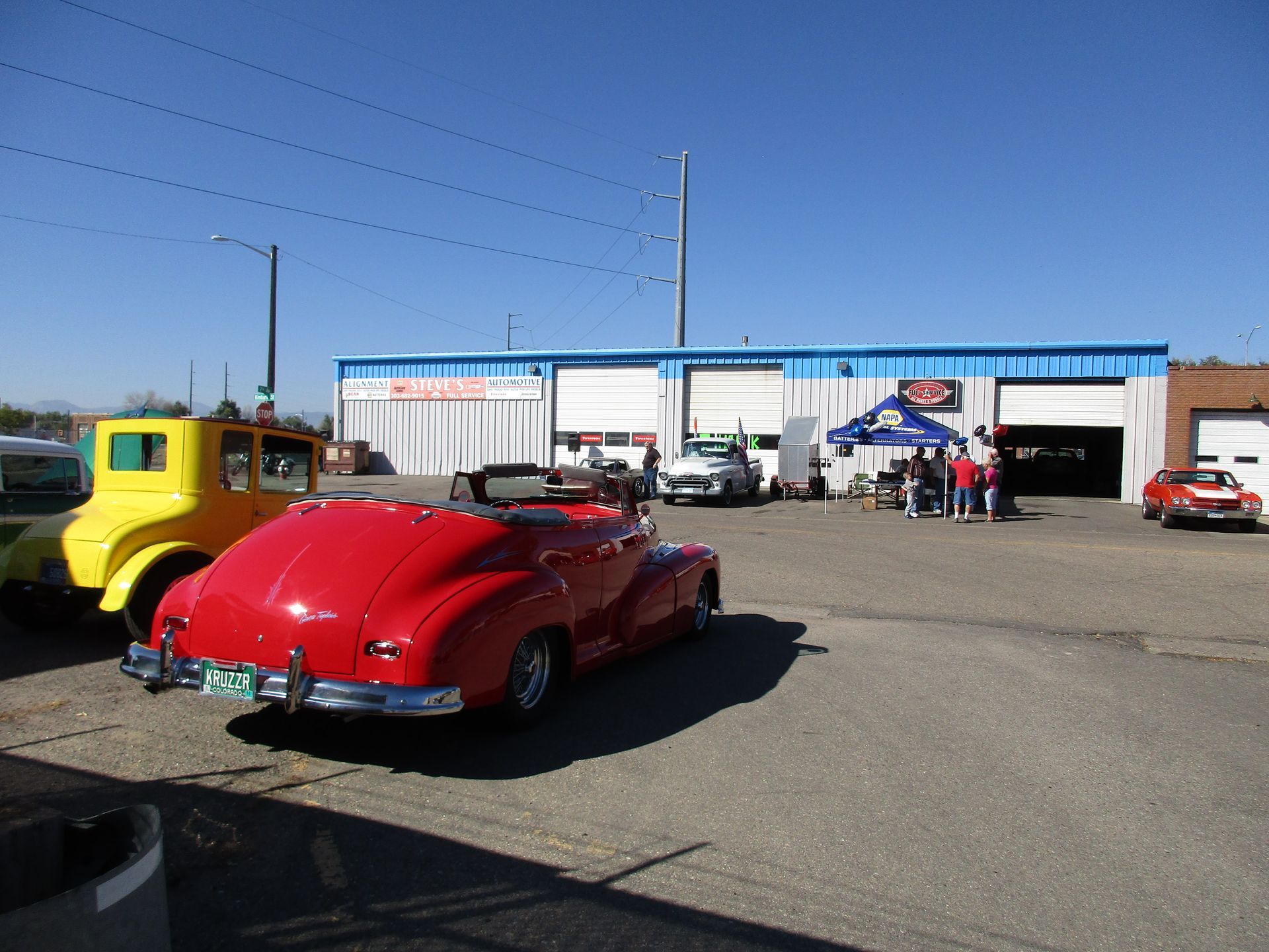Classic red convertible at an outdoor car show beside a yellow truck and blue-roofed building
